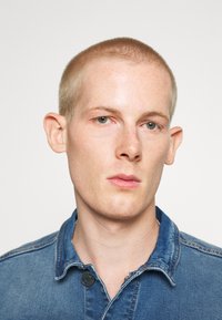 Denimskjorta med knappkrage, i en blekt blå färg och en något slitna halsringning. Nærbilds porträtt av en person.