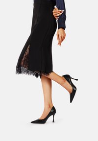 Gonna nera a matita con orlo in pizzo e spacco laterale, abbinata a tacchi in pelle nera con punta a punta e tacco a stiletto.