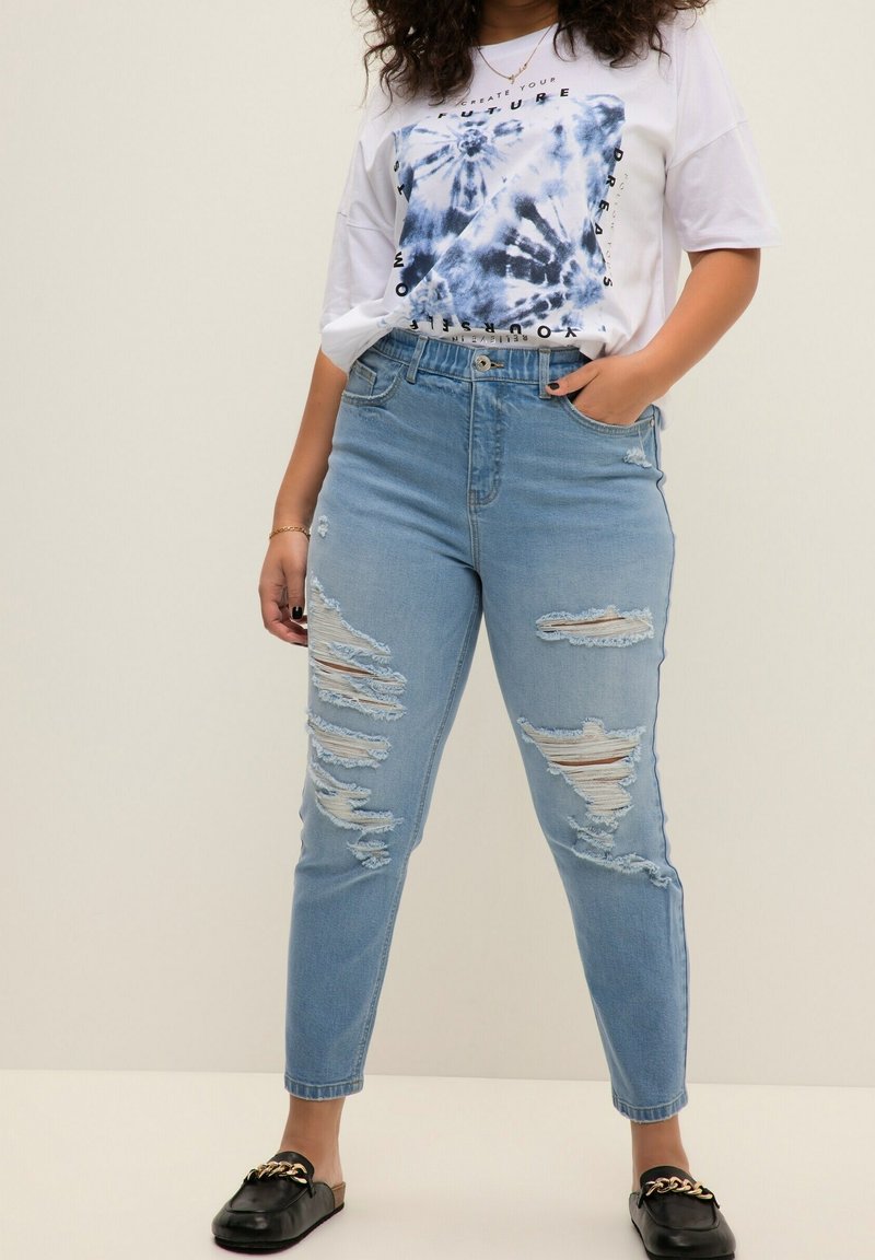 Jeans de cintura alta de color azul claro con múltiples parches desgastados y dobladillos deshilachados, combinados con una camiseta gráfica de tie-dye blanca y mocasines negros.