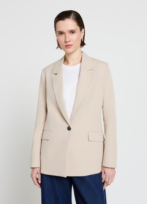 Frau mit kurzen braunen Haaren, die einen beigen Ein-Knopf-Blazer über einem weißen Hemd und dunkelblauen Hosen trägt, steht vor einem einfarbigen hellen Hintergrund.