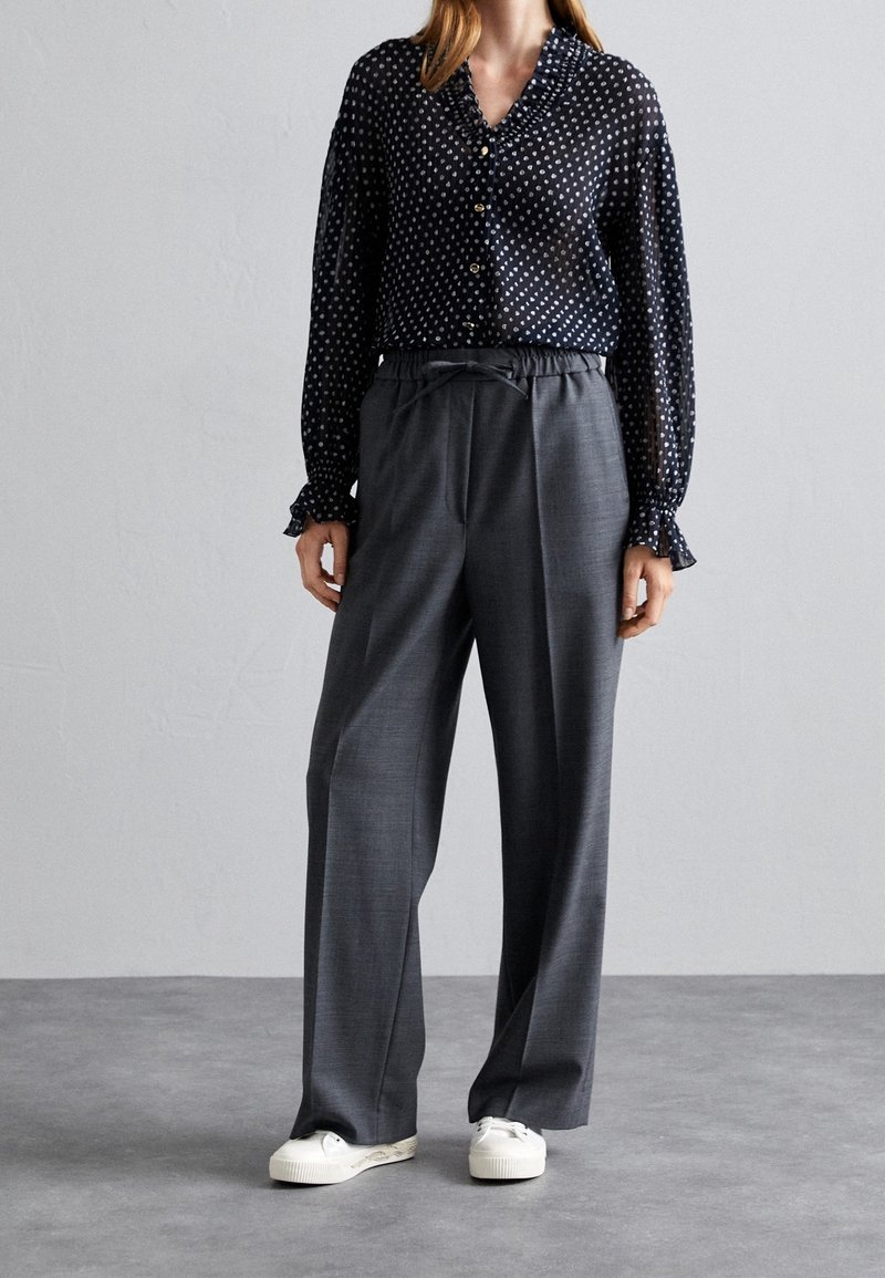 Femme portant un chemisier noir à motifs avec des poignets à volants, un pantalon gris à jambes larges avec taille à cordon et des baskets blanches, debout à l'intérieur.