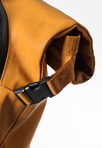 Gros plan d’un sac en cuir marron roulé avec une boucle clip en plastique noir et une sangle en tissu cousue sur fond blanc.