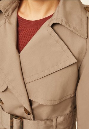 Gros plan d'une personne portant un trench-coat beige avec une ceinture sur un pull rouge foncé à côtes.
