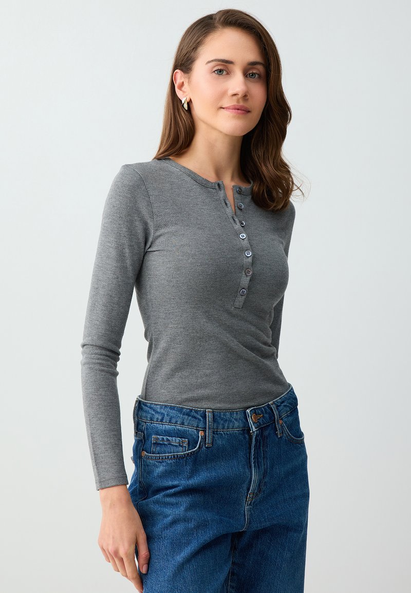 Jeune femme aux cheveux bruns ondulés portant des boucles d'oreilles dorées, un haut henley gris moulant à manches longues, et un jean bleu en denim sur un fond uni.