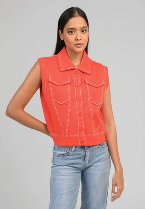 Jeune femme portant un gilet sans manches orange à boutons avec des coutures blanches et un jean bleu clair, debout devant un fond gris uni.