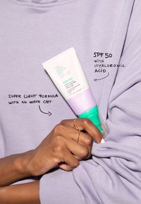Lett solkremtube med SPF 50, pastell lilla og grønn design, med tekst om hyaluronsyre og fet-fri formel.