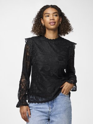 Svart bluse i blonde med en bølget halskant, lange ermer med volanger ved ermene, og blomster mønster. Kombinert med lyseblå denimjeans.