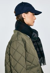 Oliwkowo zielona pikowana kurtka, granatowa czapka i ciemny, kratkowany szal z frędzlami, widziane z boku.