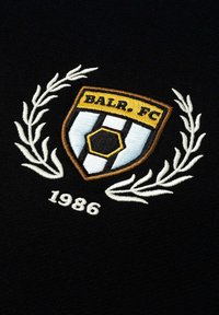 Musta kangas, jossa on brodeerattu kilpen logolla, jossa on keltaisia ja sinisiä yksityiskohtia, valkoisten laakeripensaiden ympäröimänä ja "1986" alla.