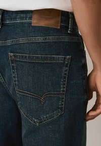 Mörkblå denimjeans med en läderlogotyp på midjebandet. Har en bakficka med gul sömnad och en klassisk design.