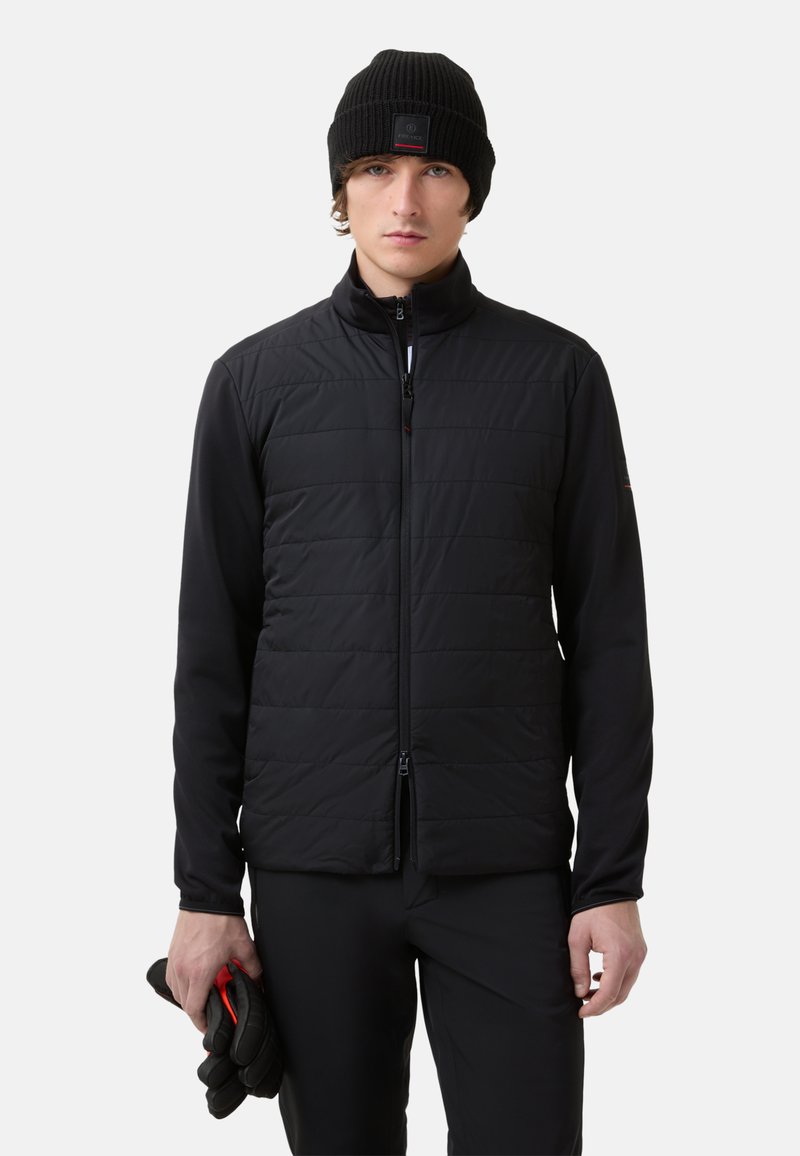 Zwarte gewatteerde vest met een quilted structuur, hoge kraag en ritssluiting; gedragen over een zwart longsleeve, gecombineerd met zwarte handschoenen.