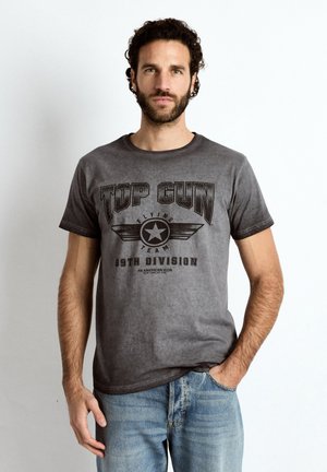 Mann mit lockigem dunklem Haar und Bart, trägt ein verblasstes graues Top Gun Grafik-T-Shirt und hellblaue Jeans, steht vor einem weißen Hintergrund.