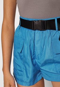 Shorts légers bleus avec une ceinture élastique, munis d'une boucle noire et de deux poches latérales pour le rangement. Texture lisse et brillante.