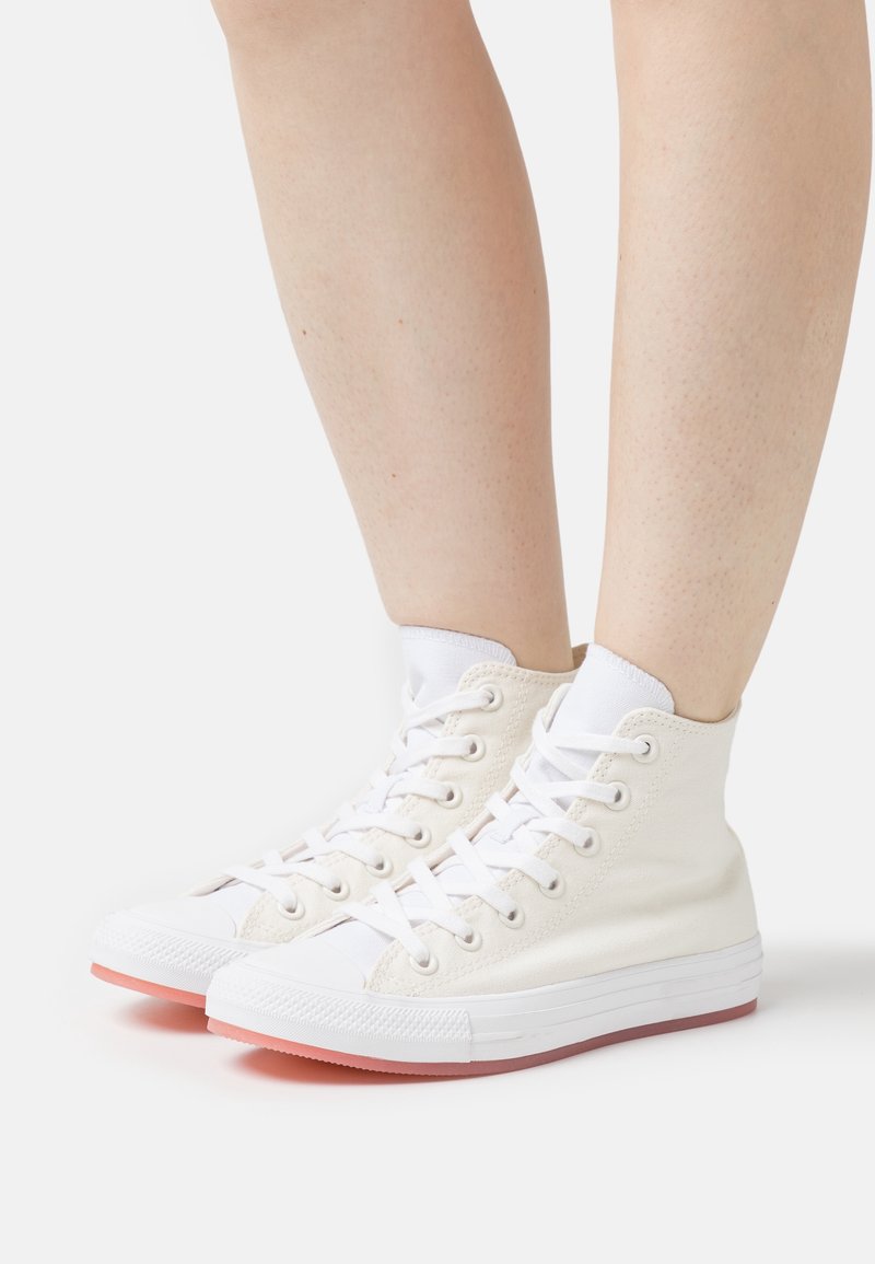 Personne portant des baskets montantes en toile blanches avec des lacets blancs et des semelles en caoutchouc rose clair sur fond blanc.