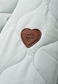 Il tessuto trapuntato azzurro chiaro presenta una patch a forma di cuore marrone con il testo in rilievo "È il momento di sbocciare". Il design cucito aggiunge texture.