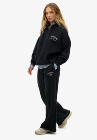 Vrouw in een zwarte zip-up hoodie en bijpassende losse broek met Superdry-logo's, beige sneakers en een gestreept shirt eronder.