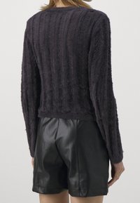 Person in einem dunklen, flauschigen Pullover und schwarzen Leder-Shorts, von hinten vor einem schlichten, hellen Hintergrund betrachtet.