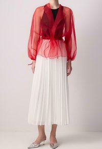 Rotes, transparentes Overlay-Top mit langen, puffigen Ärmeln und Taillenschnürung, kombiniert mit einem weißen, plissierten Midirock und silbernen spitzen Schuhen.