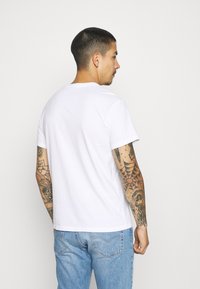 T-shirt bianco in cotone con scollo rotondo e maniche corte. Tatuaggi notevoli visibili sulle braccia. Indossato con jeans di denim azzurri chiari.