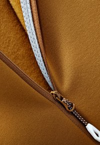 Tessuto marrone con una consistenza morbida, dotato di una cerniera grigia e dettagli marroni. La cerniera è parzialmente aperta, rivelando un rivestimento beige.