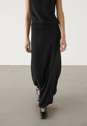 Femme marchant portant un haut noir sans manches, une longue jupe noire froncée et des chaussures noires fermées à bout avec des brides à la cheville sur un sol uni.
