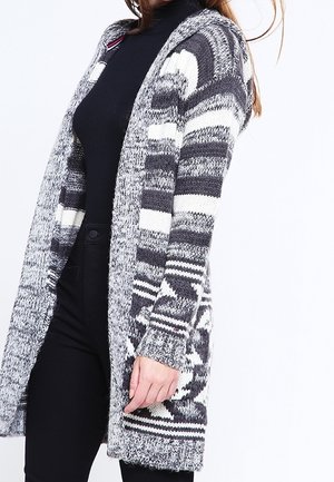 Strikket lang cardigan i sort, hvid og grå. Har et mønsterdesign med åben front og lange ærmer, lavet af tekstureret stof.