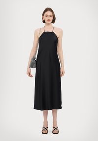 Black, sleeveless halter dress with a flowing design, smooth texture, and mid-calf length. Paired with strappy black sandals and a patterned bag.