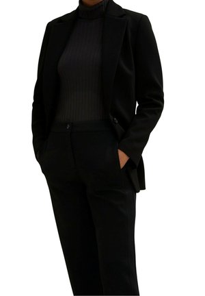 Persoon gekleed in een volledig zwarte outfit met een geribbelde coltrui, een getailleerde blazer en een broek met hoge taille, handen in de zakken, poserend tegen een witte achtergrond.