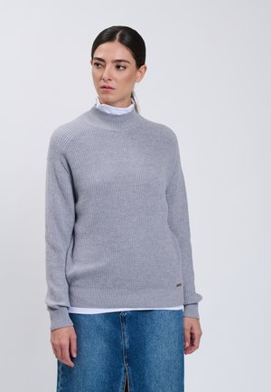 Donna con capelli scuri che indossa un maglione a maglia grigio chiaro sopra una camicia bianca e una gonna di jeans blu, che guarda di lato contro uno sfondo grigio.