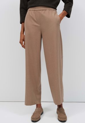 Persoon draagt beige wijde broek met elastische tailleband en bijpassende loafers, één hand in de zak, tegen een effen lichte achtergrond.