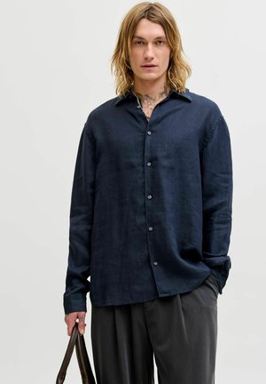 Ung person med langt blondt hår som har på seg en løs mørkeblå skjorte med knapper og grå bukser, og holder en brun skinnveske i den ene hånden.