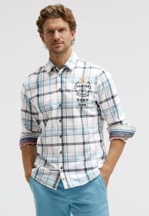 Man with curly hair wearing a light blue and white plaid shirt with rolled sleeves and light blue pants, hands in pockets.