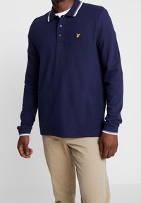 Navyn blå långärmad polo med en vit randig krage, två knappar och en liten gul logotyp på vänster bröst. Matchas med beiga byxor.