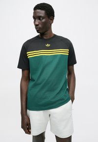 T-shirt de mangas curtas com corpo verde escuro, mangas pretas e riscas amarelas no peito, com um pequeno logótipo acima.