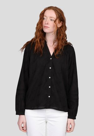 Blouse noire, légère, avec un décolleté en V, des manches longues, une fermeture à boutons et un subtil motif floral embossé. Les boutons blancs contrastent avec le tissu.