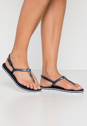 Pieds féminins portant des sandales plates noires et blanches avec des fines lanières sur une surface et un fond blancs.