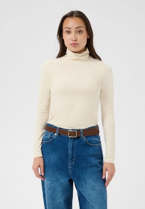 Jeune femme aux longs cheveux foncés portant un col roulé crème, un jean bleu taille haute et une ceinture marron, debout devant un fond uni.
