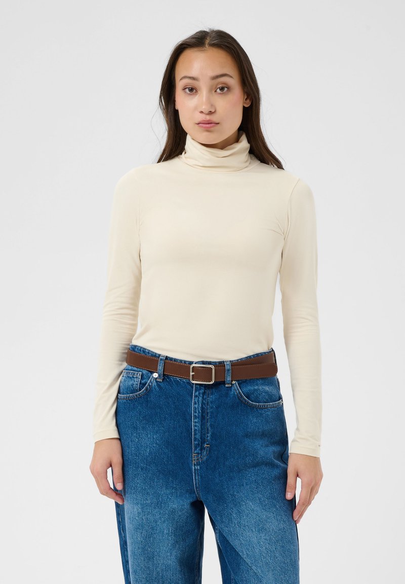 Jeune femme aux longs cheveux foncés portant un col roulé crème, un jean bleu taille haute et une ceinture marron, debout devant un fond uni.