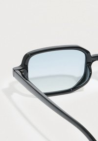 Μαύρα ορθογώνια γυαλιά ηλίου με λεπτούς βραχίονες και ελαφρώς βαμμένους μπλε φακούς, που ακουμπούν σε λευκή επιφάνεια.