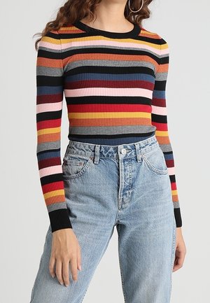 Femme portant un pull côtelé ajusté à manches longues avec des rayures horizontales colorées et un jean bleu clair taille haute.