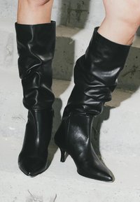 Picioare purtând cizme de piele negre, slouchy, până la mijlocul gambelor, cu vârfuri ascuțite și tocuri kitten, stând pe trepte de beton.