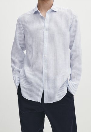 Man wearing a long-sleeve white shirt with thin blue vertical stripes, tucked loosely into black pants with hands in pockets.