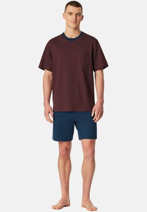 Mann steht barfuß, trägt ein dunkelrotes kurzärmeliges Hemd mit Tasche und marineblaue Shorts vor einem schlichten weißen Hintergrund.