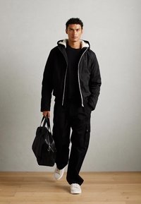 Giacca nera con zip e cappuccio bordato di bianco, abbinata a una maglietta nera a maniche lunghe e pantaloni cargo. Il modello tiene una borsa in pelle nera.