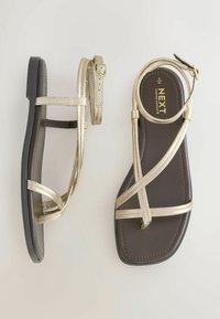 Par af damesandaler med flade såler, tynde metallisk gyldne remme og mørkebrune fodsenge, den ene sandal set oppefra og den anden fra siden.