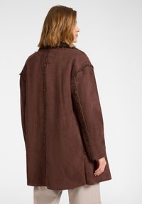 Manteau en faux daim marron avec une coupe décontractée, des bords effilochés et une doublure texturée. Il présente un design simple et une forme doucement structurée.