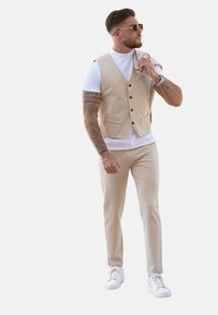 Gilet beige par-dessus un t-shirt blanc, pantalon beige et baskets blanches. Comprend des boutons sur le gilet, des lignes épurées et des tatouages sur les bras.
