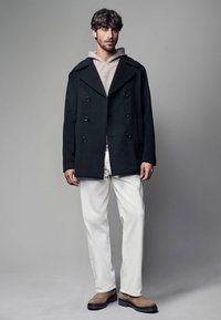 Schwarzer Peacoat mit doppelt geknöpften Knöpfen über einem beigen Hoodie, kombiniert mit weißen Hosen und braunen Wildlederstiefeln. Glatte Textur, taillierte Passform.