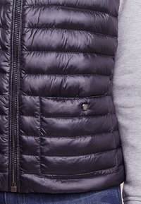 Navy puffer vest featuring quilted sections, front zipper, and a snap detail, made from glossy, lightweight material. Textured gray garment underneath.