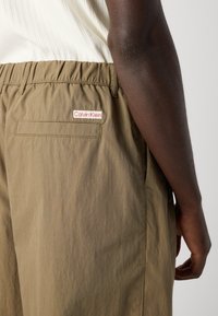 Close-up of a person wearing khaki Calvin Klein pants with elastic waistband and back pocket, paired with a white ribbed top.